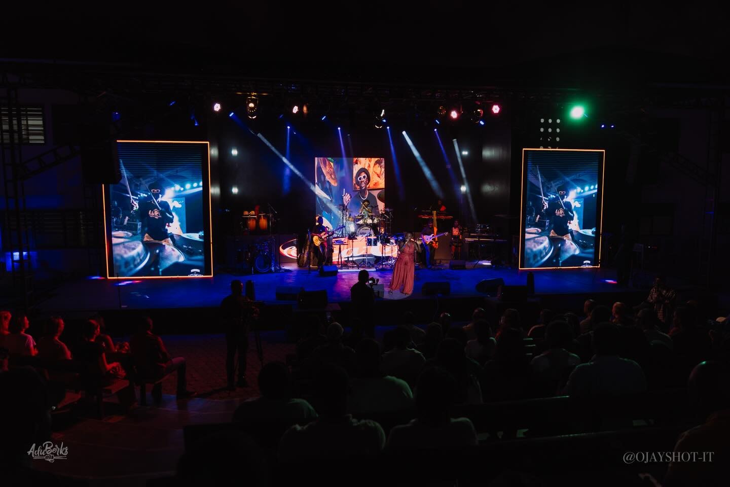 Dark stage lit in blue with performers and side screens